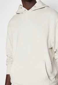 Felpa con cappuccio beige chiaro con tasca a marsupio, cappuccio regolabile con coulisse e bottoni a pressione. Realizzata in tessuto morbido e strutturato, con vestibilità comoda.