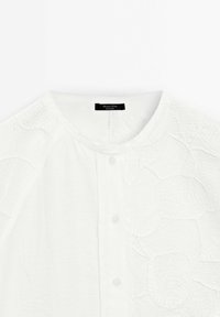Blusa bianca a pulsante con scollatura rotonda e dettagli in pizzo floreale testurizzato sulle spalle e sulla parte superiore del petto, etichetta visibile al colletto.