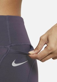 Les leggings violets Nike présentent un design épuré avec des coutures plates et une petite poche, agrémentés d'un logo argenté sur le côté.