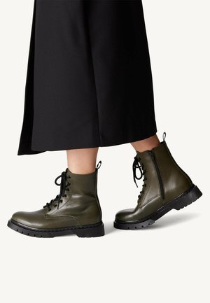 Bottes de cheville en cuir vert avec des lacets noirs, zip sur le côté, semelle texturée et bout arrondi. Portées avec une jupe noire, mettant en valeur le design des bottes.