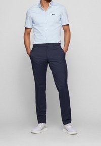 Light blue short-sleeve button-up shirt with black logo, paired with dark navy dress pants and white sneakers, standing posture, hands on hips.