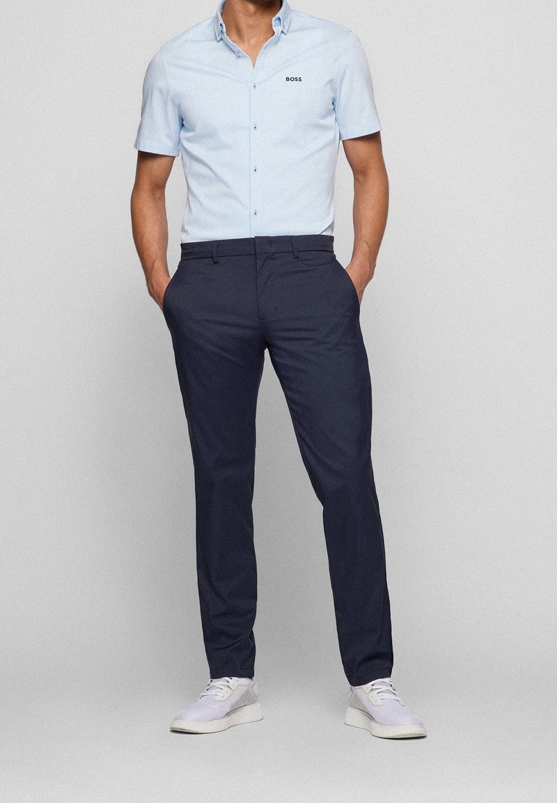 Light blue short-sleeve button-up shirt with black logo, paired with dark navy dress pants and white sneakers, standing posture, hands on hips.