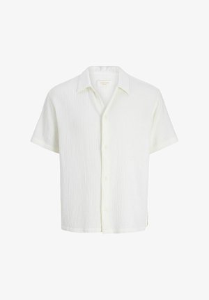 Garçon portant une casquette rose pâle, une chemise blanche à manches courtes texturée avec boutons, un short noir, des chaussettes blanches et des chaussures beiges, regardant sur le côté.