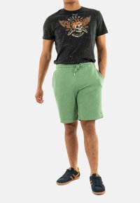 Pantalones cortos de algodón verde con cinturilla elástica, combinados con una camiseta gráfica negra que presenta un diseño de calavera y alas. Zapatillas negras y naranjas.