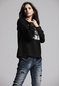 Black hoodie with a graphic print, cinched sleeves, and a relaxed fit, paired with distressed blue jeans featuring rips and a faded wash.