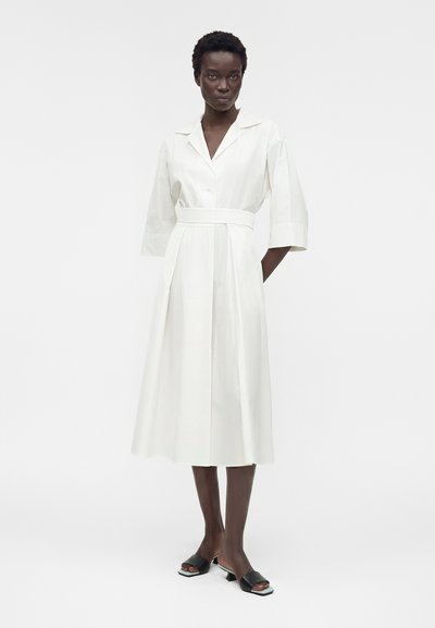 Γυναίκα στέκεται φορώντας ένα λευκό midi φόρεμα με γιακά, μανίκια τριών τετάρτων, ζώνη στη μέση και μαύρα ψηλοτάκουνα σανδάλια με ανοιχτά δάχτυλα.