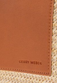 Skórzana etykieta w kolorze brązowym, przyszyta do plecionego słomianego tła, z wytłoczonym napisem "GERRY WEBER" wielkimi literami. Brak innych szczegółów.