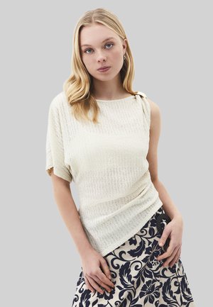 Jeune femme aux cheveux blonds portant un haut tricoté texturé blanc cassé avec une manche et une jupe à motif floral noir et blanc.