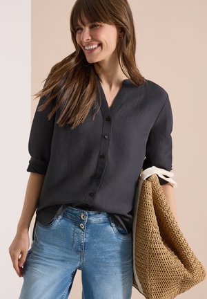 Femme souriante, portant une chemise noire boutonnée avec les manches retroussées et un jean bleu clair, tenant un grand sac cabas tressé beige.