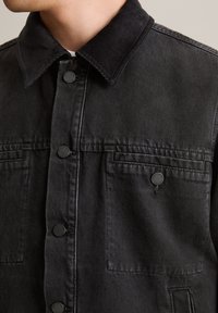 Zwarte spijkerjack met een corduroy kraag, drukknopen, twee borstzakken en een licht getextureerde afwerking.