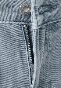 Svetlomodrá rifľovina s viditeľným strieborným zipsom a kovovou gombíkovou nášivkou s nápisom "MNG DENIM." Zreteľný textúrovaný vzor tkaniny.