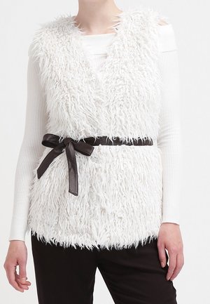 Personne portant un gilet en fausse fourrure blanche texturée avec une ceinture noire sur un haut blanc à manches longues et un pantalon noir, vue des épaules aux hanches.