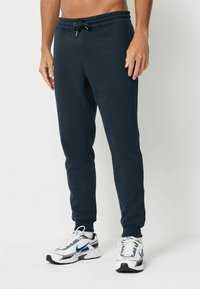 Tumši zili sporta bikses no mīksta materiāla ar elastīgu jostu, regulējamu auklu un sašaurinātiem piedurkņu galiem, apvienotas ar baltām un pelēkām kedām.