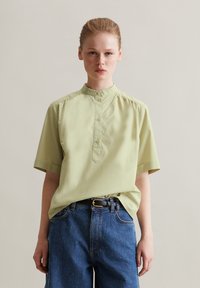 Camicia verde chiaro a maniche corte con colletto alla cinese, apertura frontale con bottoni e dettaglio arricciato sulle spalle, indossata con pantaloni in denim blu.