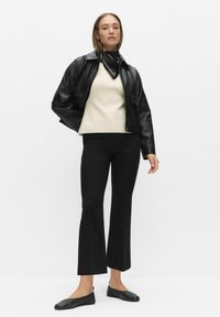 Zwarte leren jas, beige trui, zwarte uitlopende broek en zwarte ballerina's. De jas heeft voorzakken en een kraag; een sjaal zorgt voor een contrast.