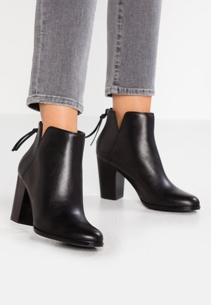 Schwarze Leder-Stiefeletten mit klobigen Absätzen, getragen zu grauen gekürzten Jeans auf weißem Boden und Hintergrund.