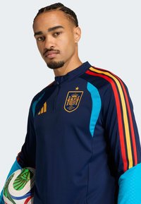 Casaco de treino da seleção nacional de Espanha em azul-marinho, com riscas vermelhas, amarelas e azuis, fecho até ao meio da altura do peito e emblema no peito. Bola segurada na mão.