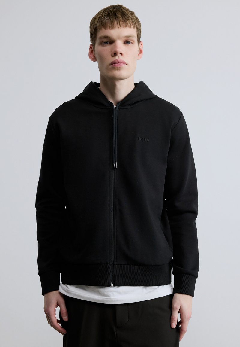 Črni pulover s kapuco z zadrgo, narejen iz mehke bombažne tkanine. Ima kapuco z zavezovanjem, rebraste manšete in subtilno logotip na prsih. Gladek otip.