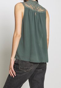 Top verde senza maniche con dettagli in pizzo sul giromanica, apertura a forma di chiave sul retro, e tessuto fluido, abbinato a jeans scuri.