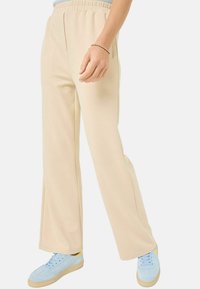 Beige, stretchy, elastic-waist pants with two side pockets and a relaxed fit, paired with light blue sneakers featuring a yellow accent.