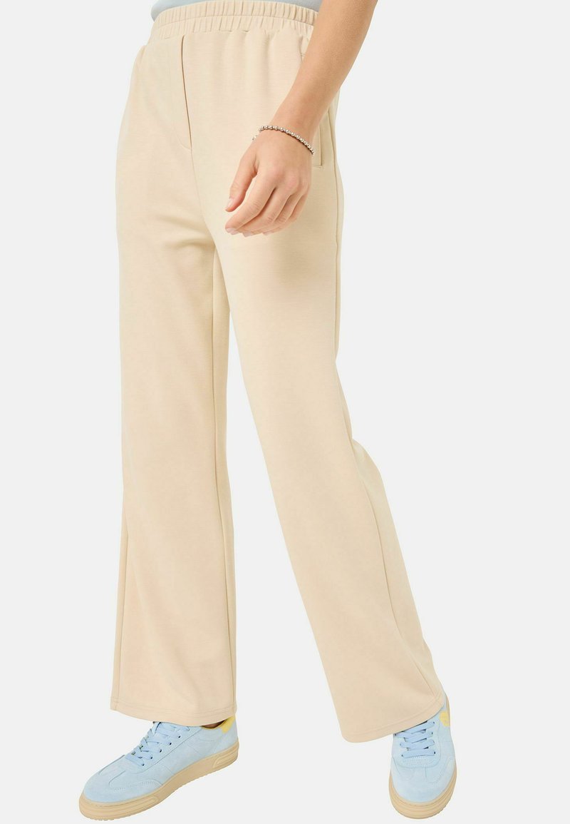 Beige, stretchy, elastic-waist pants with two side pockets and a relaxed fit, paired with light blue sneakers featuring a yellow accent.