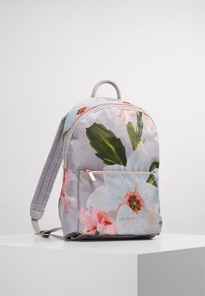 Sac à dos gris clair avec un grand imprimé floral blanc et rose, poche avant zippée et bretelles rembourrées sur une surface blanche.