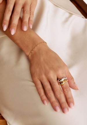 Goldkettchen-Armband und ein silber-gold verflochtener Ring an einer Hand mit hellrosa lackierten Nägeln, die auf einer beigen Stoffoberfläche ruht.