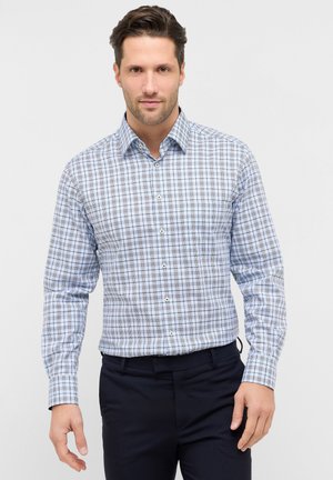 Mężczyzna ubrany w jasnoniebieską i szarą kratowaną koszulę z długim rękawem oraz czarne spodnie, stojący na tle jednolitego jasnego tła.