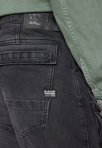 Svart denimjeans har en strukturerad yta, rak skärning och synlig sömnad. En läderetikett visar "G-Star RAW." En grön skjorta syns ovanför.