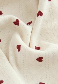 Kreemivärvi kudum, millel on tekstuurne muster ja hajutatud punased südamed. Pehme ja paindlik materjal, millel on sile pind.