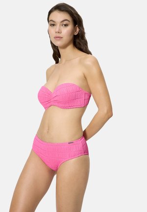 Joven mujer con un bikini rosa texturizado sin tirantes y braguitas de talle alto a juego, de pie frente a un fondo claro y liso.