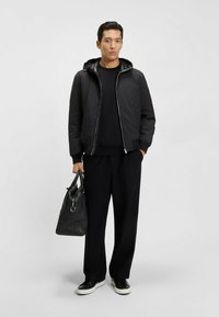 Man som bär svart huvjacka, svarta byxor och svarta sneakers med vita sulor, håller en svart läderhandväska med ena handen.