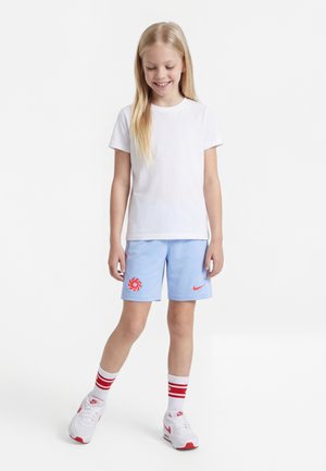 Tyttö seisoo ja hymyilee, hänellä on valkoinen t-paita, vaaleansiniset shortsit punaisilla logoilla, valkoiset sukat punaisilla raidoilla ja valkoiset lenkkarit.