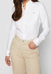 Hvid skjorte med knapper i bomuld med en lille logodetalje. Sammen med lysebeige high-waisted jeans med standardlommer og syninger.