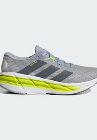 Zapatilla deportiva de color gris claro con un material superior texturizado, rayas negras y detalles en amarillo brillante en la suela, que cuenta con un diseño de entresuela acolchada.