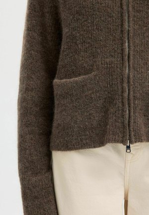 Gros plan sur une personne portant un pull zippé brun en tissu pelucheux avec une poche avant et un pantalon de couleur crème.