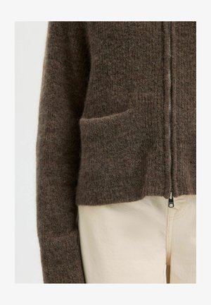 Gros plan sur une personne portant un pull zippé brun en tissu pelucheux avec une poche avant et un pantalon de couleur crème.