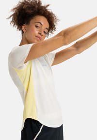 Camisa deportiva blanca con paneles de malla amarillos a los lados, mangas cortas y un ajuste relajado. Presenta un pequeño logo en la parte frontal. Se llevan pantalones cortos negros.