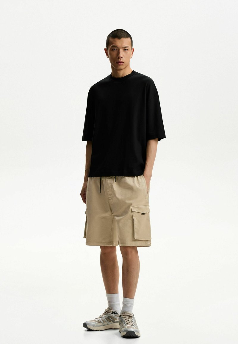 Tricou oversized negru cu mâneci scurte, purtat cu pantaloni scurți cargo bej, dotati cu buzunare laterale. Tenis gri completează ținuta.