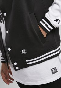 Zwart-witte varsityjack met een drukknopenfront, contrasterende mouwen, gestreepte manchetten en zak, met logo patches aan de voorkant.