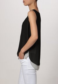 Top senza maniche nero con un design fluido, caratterizzato da uno strato posteriore grigio chiaro leggermente più lungo, indossato con pantaloni bianchi, tessuto di texture liscia.