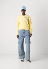 Felpa gialla con logo, pantaloni cargo in denim blu larghi e sandali azzurri chiari. Modello che indossa un berretto abbinato, in piedi su uno sfondo bianco.