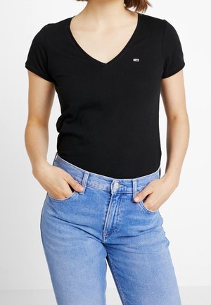 Vrouw die een zwart V-hals T-shirt met korte mouwen draagt, met een klein logo op de borst en lichtblauwe high-waisted jeans, handen in de zakken.