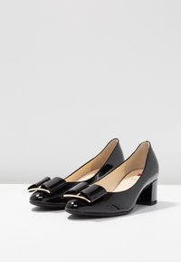 Svarta lackläder pumps med låga klackar, rundade tår, blockklackar och dekorativa svarta rosetter med guldkant på en vit yta.