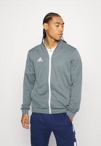 Grå atletisk jakke med lynlås foran, ribbede manchetter og sidelommer. Har det hvide Adidas-logo på venstre bryst. Bæres over en hvid skjorte.