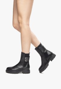 Black leather ankle boots with a chunky sole, elastic side panels, and metallic accent detailing, featuring a side zipper for easy wear.