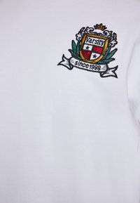 Hvid bomulds t-shirt med et broderet våbenskjold med teksten "Varsity" og "siden 1998", der inkluderer grønne og røde accenter.