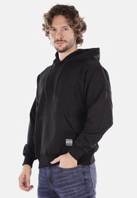 Felpa nera con cappuccio, dotata di tasca a marsupio e cordini, realizzata in tessuto morbido, indossata da un modello con jeans scuri.