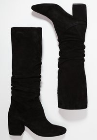 Svarta mockaskinnsstövlar i knähöjd med en slapp design, spetsig tå och kraftig klack. Texturen är mjuk och tyget verkar slätt.