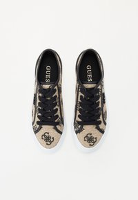 Scarpe da ginnastica in tela beige con motivo e logo neri, dotate di lacci neri e suola in gomma bianca. Texture liscia e design classico.